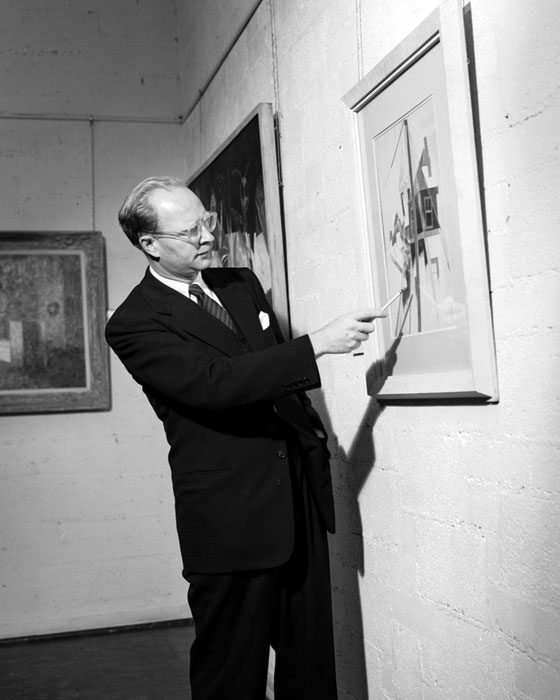 A black-and-white photo of a man wearing glasses and a suit. He is pointing at a piece of framed artwork on the wall.