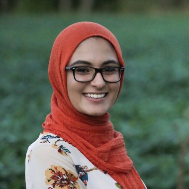 A woman with olive skin smiles at the camera. She wears dark-rimmed glasses, a floral print top, and an orange-red hijab.