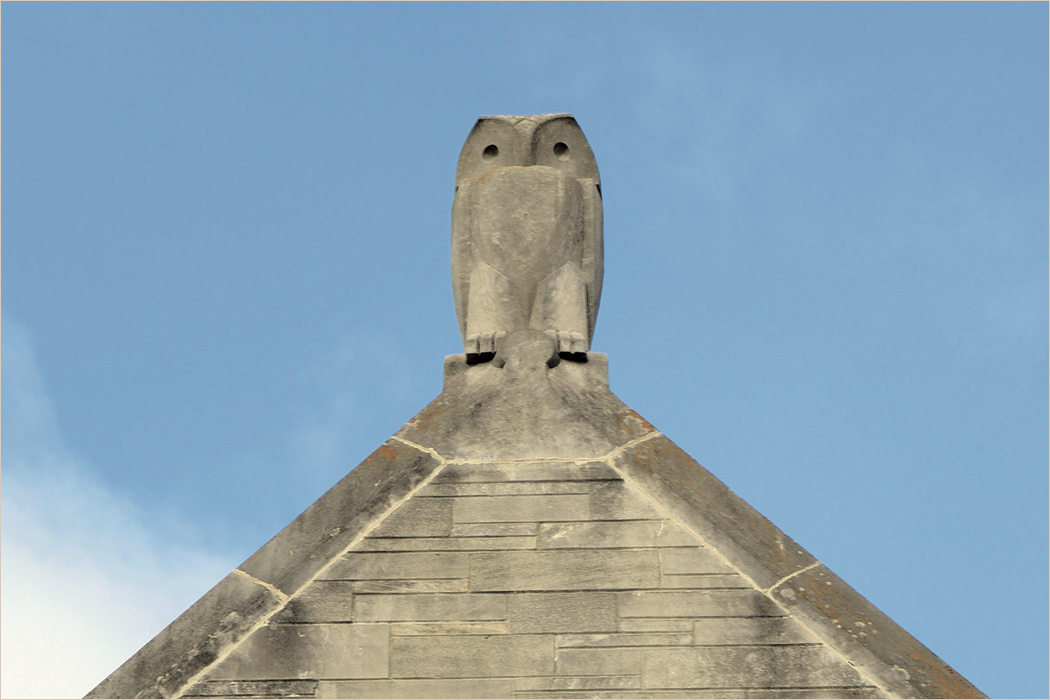 A limestone sculpture of an owl sitting on the peak of a roof.